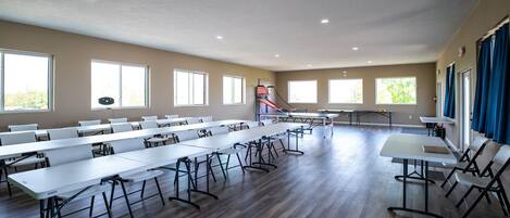 A spacious room with multiple tables and chairs, large windows, and a basketball hoop.