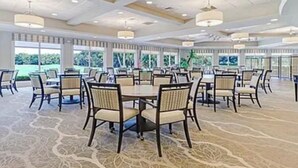 A spacious banquet hall with round tables and chairs, large windows offering a view of greenery, and a patterned carpet. - Gated FoxFire Naples First-Floor/End-unit Amazing Water/Course View 27-Hole Golf (Naples)