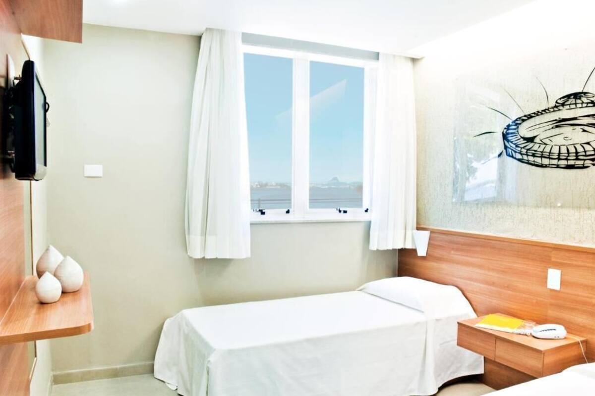 A hotel room with a single bed, a bedside table with a lamp, a window with curtains, and a view of the sea.
