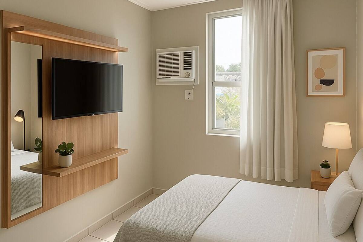 A hotel room with a large bed, a mounted TV, a window with curtains, a bedside lamp, and a small plant on the nightstand.