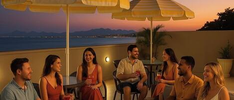 A group of people seated around a table on a terrace with umbrellas, overlooking a body of water and mountains.