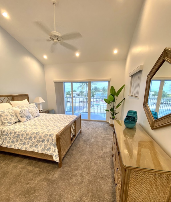 A bedroom with a bed, a dresser, a ceiling fan, and a large window with a view of palm trees and a swimming pool.