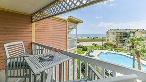 A balcony with a table, chairs, and a magazine. There is a pool with palm trees and a view of a beach and buildings.