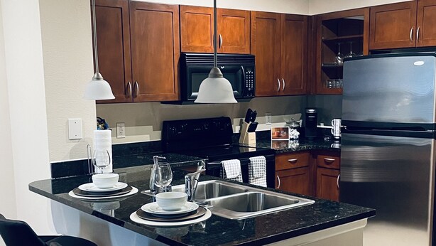 Una cucina moderna con mobili in legno, un frigorifero in acciaio inossidabile, un forno a microonde e un piano di lavoro nero. Sono presenti due sgabelli da bar e un cestino.
