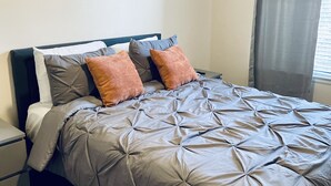 Un letto rifatto con cura, coperto da una trapunta grigia e con due cuscini arancioni, un comodino con una lampada e un quadro appeso al muro.