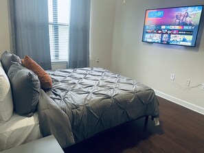 Ein Schlafzimmer mit einem Bett, einem an der Wand montierten Fernseher und einem Fenster mit Vorhängen.