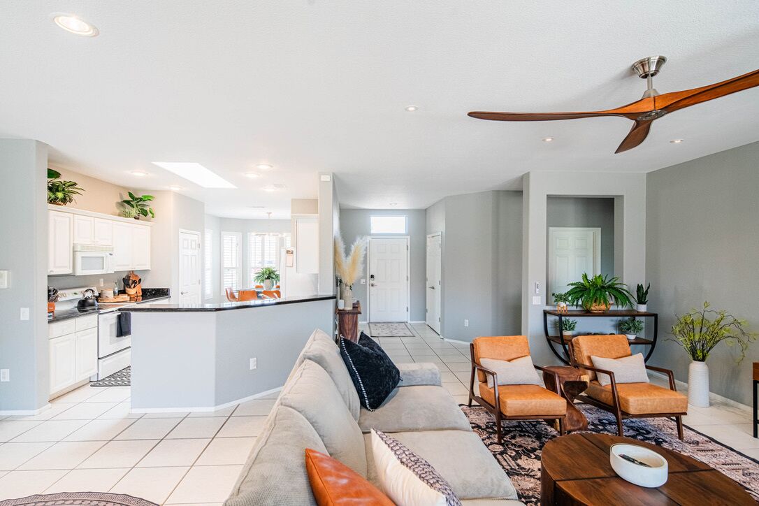 Un vaste espace de vie comprenant un canapé, une table basse et des fauteuils, une cuisine avec des meubles blancs et un ventilateur de plafond.