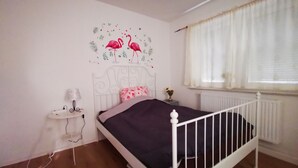 A bedroom with a white metal bed frame, a purple bedspread, a wall decal of flamingos, a bedside table with a lamp, and a window with white curtains.
