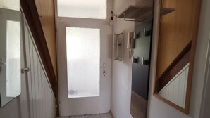 A narrow hallway with a door leading to a room. The hallway has a tiled floor with a diamond pattern. There is a light fixture on the wall and a small shelf above it.