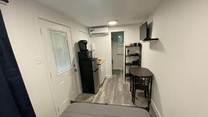 A compact hotel room with a bed, a small kitchenette, a dining table with chairs, and a TV mounted on the wall.