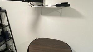 A room with a flat-screen TV mounted on the wall, a small table with a chair, and a shelf with various items.