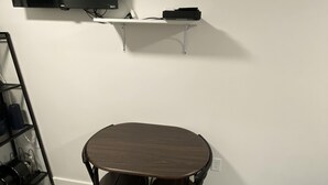 A room with a flat-screen TV mounted on the wall, a small white shelf below it, and a dark wooden table with four chairs.