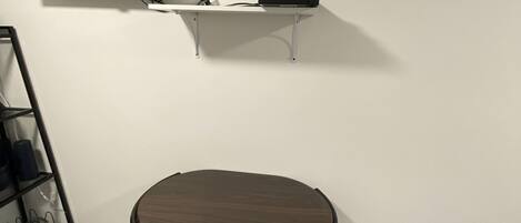 A room with a flat-screen TV mounted on the wall, a small white shelf below it, and a dark wooden table with four chairs.