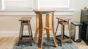 Une pièce avec une table en bois et deux tabourets, un tapis gris et une poubelle.