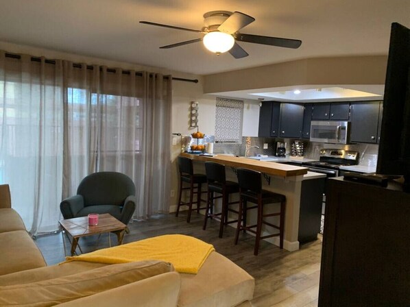 Una sala de estar moderna con un sofá, una mesita y un sillón. Hay una zona de cocina con una barra y una mesa de comedor.