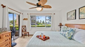 A bedroom with a large bed, a ceiling fan, a window with a view of greenery, a wooden dresser, and a bedside table with a lamp.