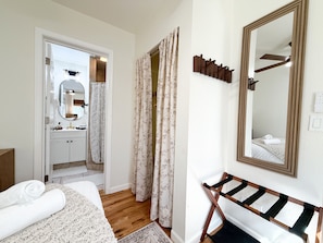 A bedroom with a bed, a wooden bench, a mirror, and a bathroom with a sink and shower.