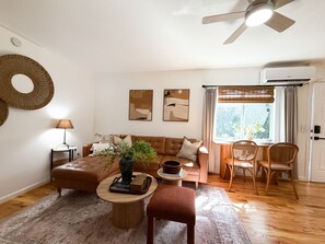 Una sala de estar con un sofá de cuero marrón, una mesa redonda, una silla de mimbre y una ventana con cortinas.