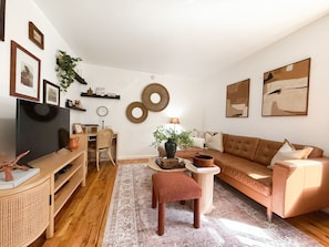 Una sala de estar con un sofá marrón, una mesa de centro de madera, una silla de mimbre, un televisor y decoraciones en la pared, incluyendo cuadros enmarcados y un adorno de pared circular.