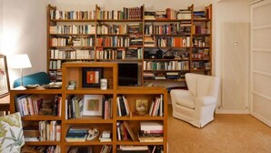 A cozy living room with a large wooden bookshelf filled with books, a comfortable white armchair, and a small table with a lamp.