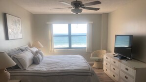 A bedroom with a bed, a television, a window with a view of the ocean, a ceiling fan, and a wicker chair. - Paradise in Ponce - Oceanfront 2BR Condo (Ponce Inlet)