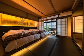 A traditional Japanese-style hotel room with tatami flooring, a large bed with white and brown bedding, a shoji screen door, and a view of greenery outside.