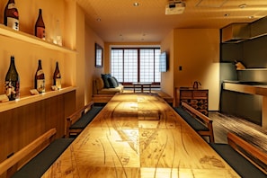 Ein langer Holztisch mit aufwendigen Verzierungen, eine hölzerne Bank und eine Couch mit Kissen. Regale mit Flaschen und ein Fernseher an der Wand sind ebenfalls vorhanden.