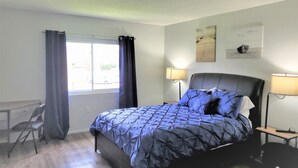 Une chambre à coucher avec un lit, deux lampes de chevet, une fenêtre avec des rideaux, une chaise et des œuvres d’art accrochées au mur.