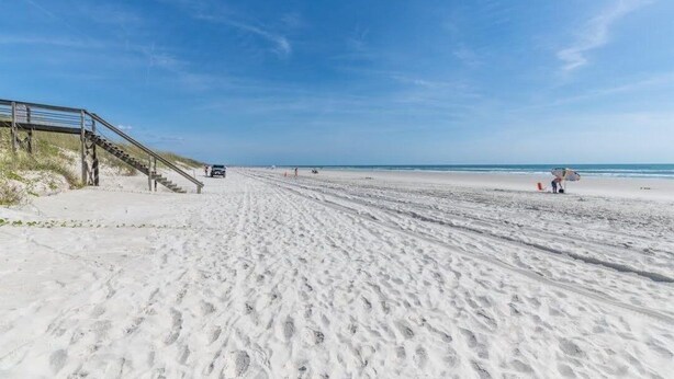 Une plage de sable avec un escalier descendant vers l’eau, quelques personnes et un ciel bleu dégagé.