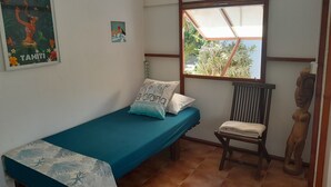 Una habitación pequeña con cama individual, colcha color aguamarina, una silla de madera, una ventana con vistas a zonas verdes y un cuadro enmarcado de una escena tropical.