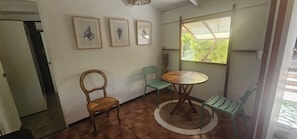 A room with a wooden table, two chairs, a window with a view of greenery, and three framed pictures on the wall.