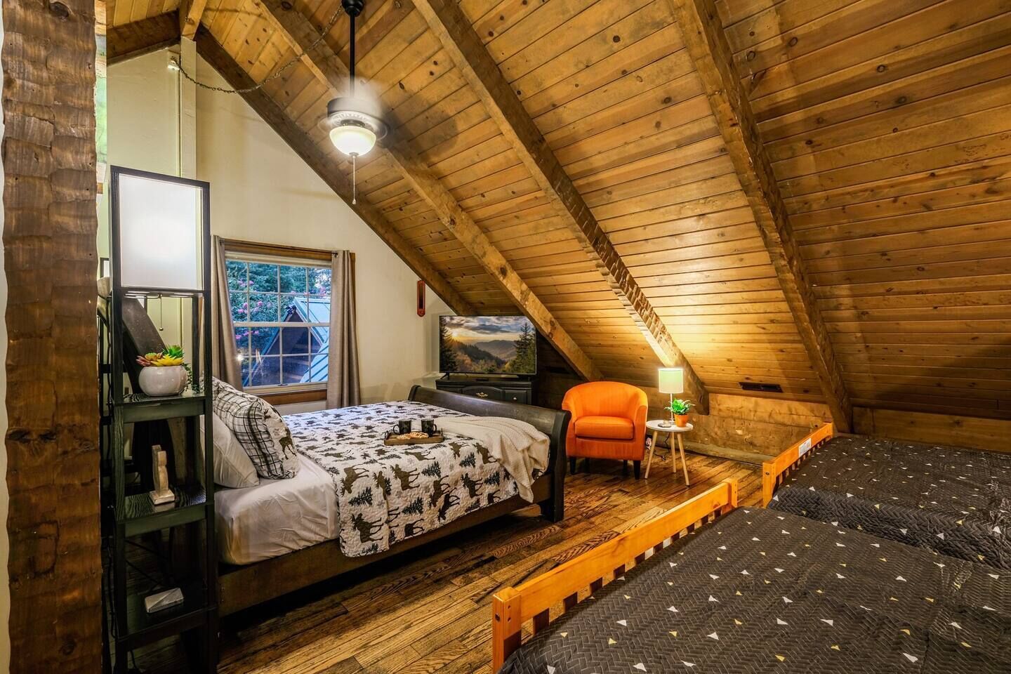A cozy attic bedroom with a bed, a chair, a TV, and a window overlooking a scenic view.