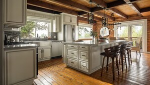 A kitchen with white cabinets, a large island with bar stools, wooden beams on the ceiling, and a view of the outdoors. - Chateau Syrah by AvantStay | Picturesque Estate w/ Pool, Pickleball, Hot Tub (Temecula)
