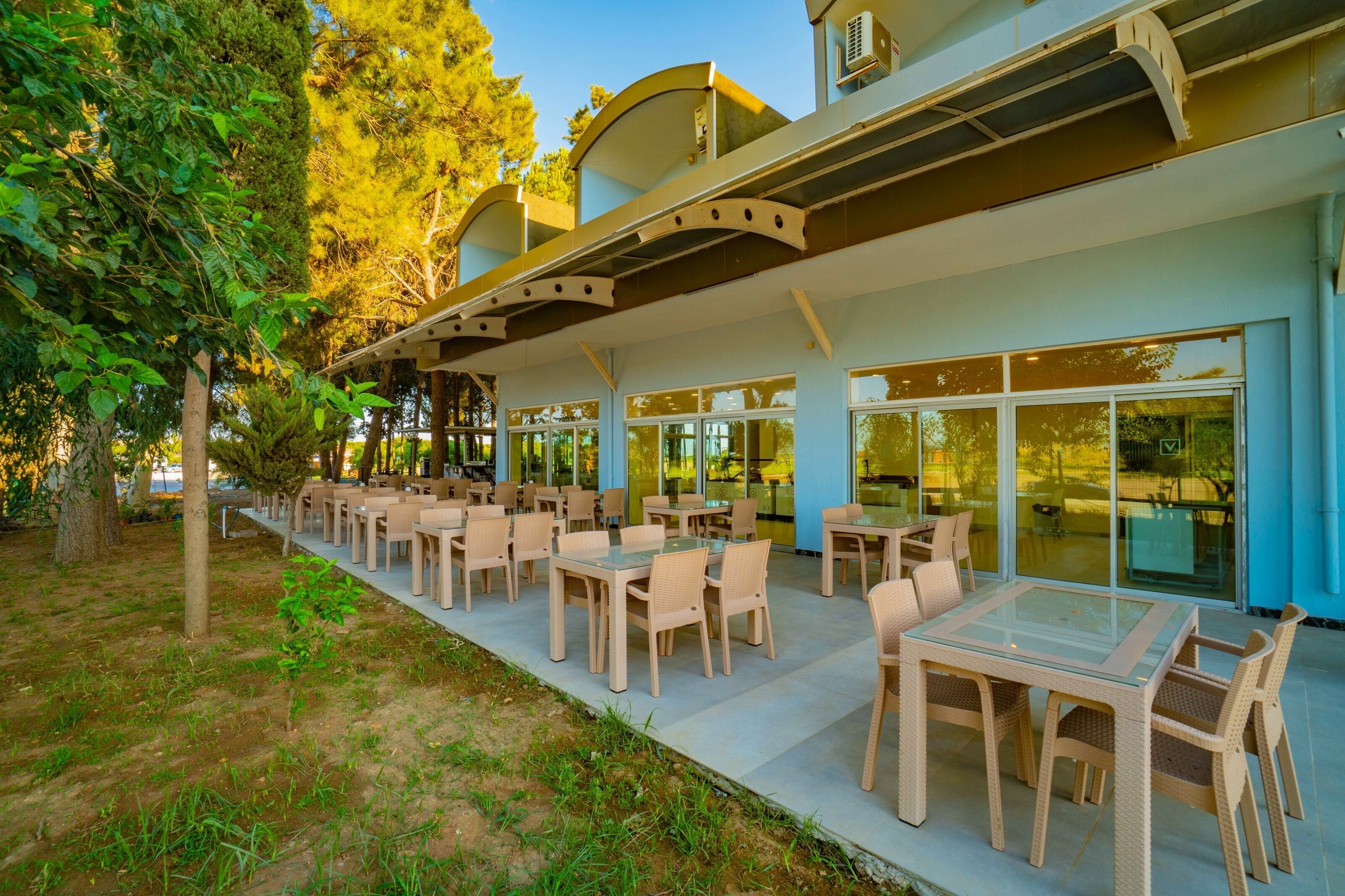 Un espace extérieur aménagé avec des tables et des chaises, entouré d’arbres et bordé d’un bâtiment doté de grandes fenêtres.