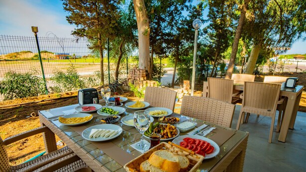 Un espace de restauration en plein air avec des chaises en osier et une table en verre, dressée avec des assiettes de nourriture, un panier de pain et une bouteille d’eau.