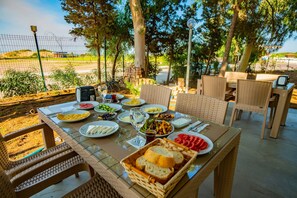 An outdoor dining area with wicker chairs and a glass table set with plates of food, a basket of bread, and a bottle of water. - Prenses Sealine Beach Hotel (Serik)