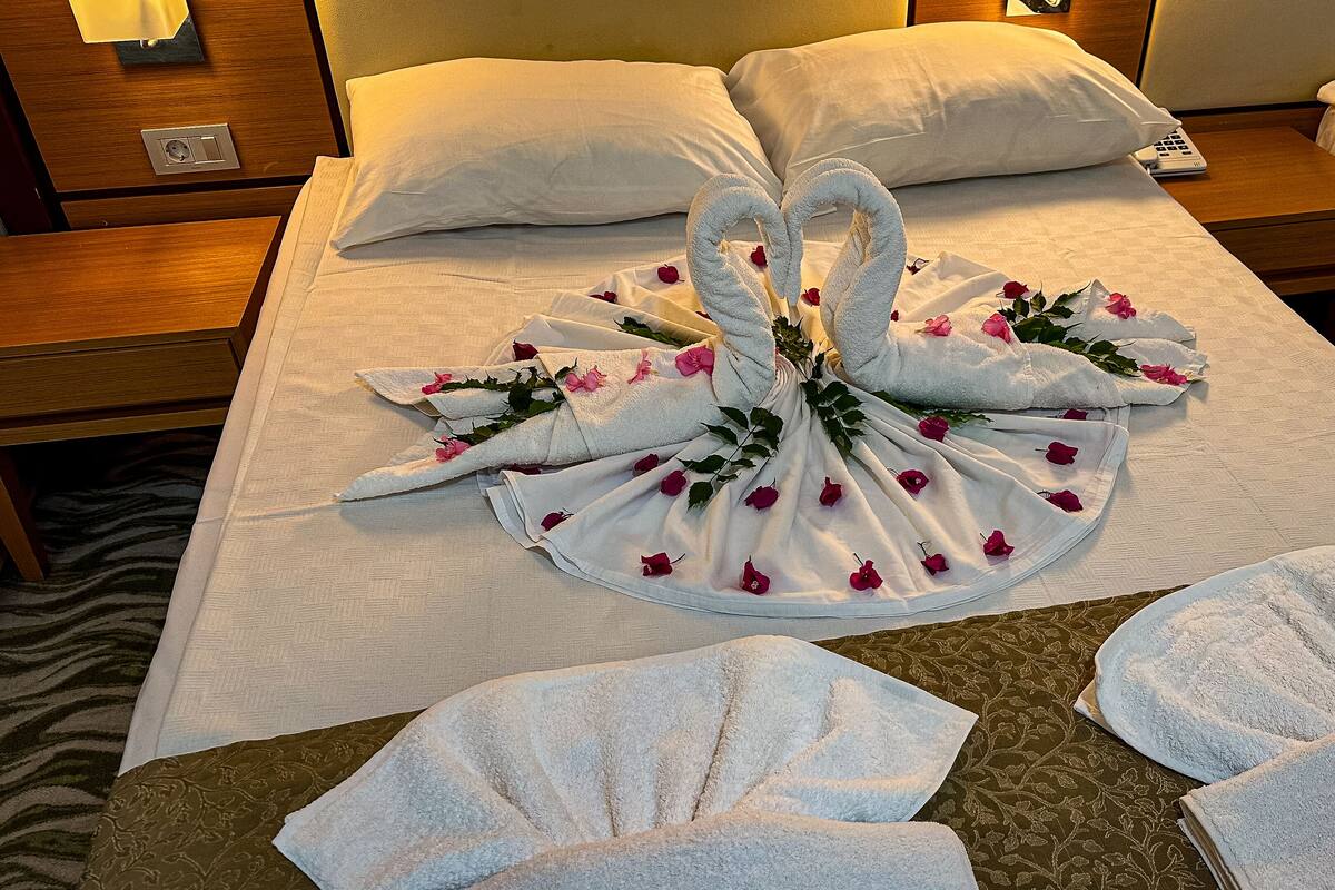 A neatly made bed with a towel arrangement in the shape of a heart, featuring rose petals and greenery.