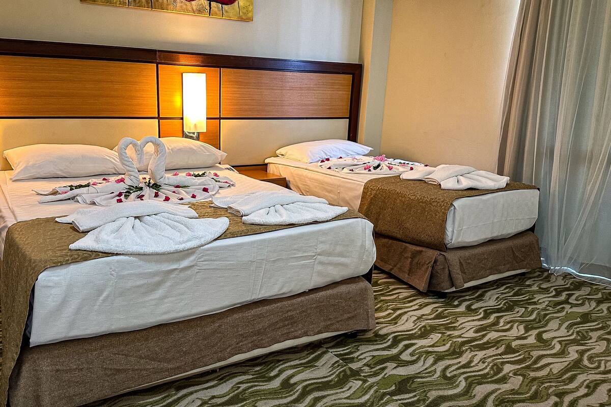 A hotel room with two beds, each with a brown bedspread, white pillows, and towels arranged in a decorative manner. There are wall-mounted lamps on either side of the headboard, and a painting above the beds.