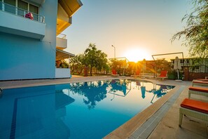 A swimming pool with clear water reflecting trees and a sunset. There are lounge chairs and a covered area with umbrellas. - Prenses Sealine Beach Hotel (Serik)
