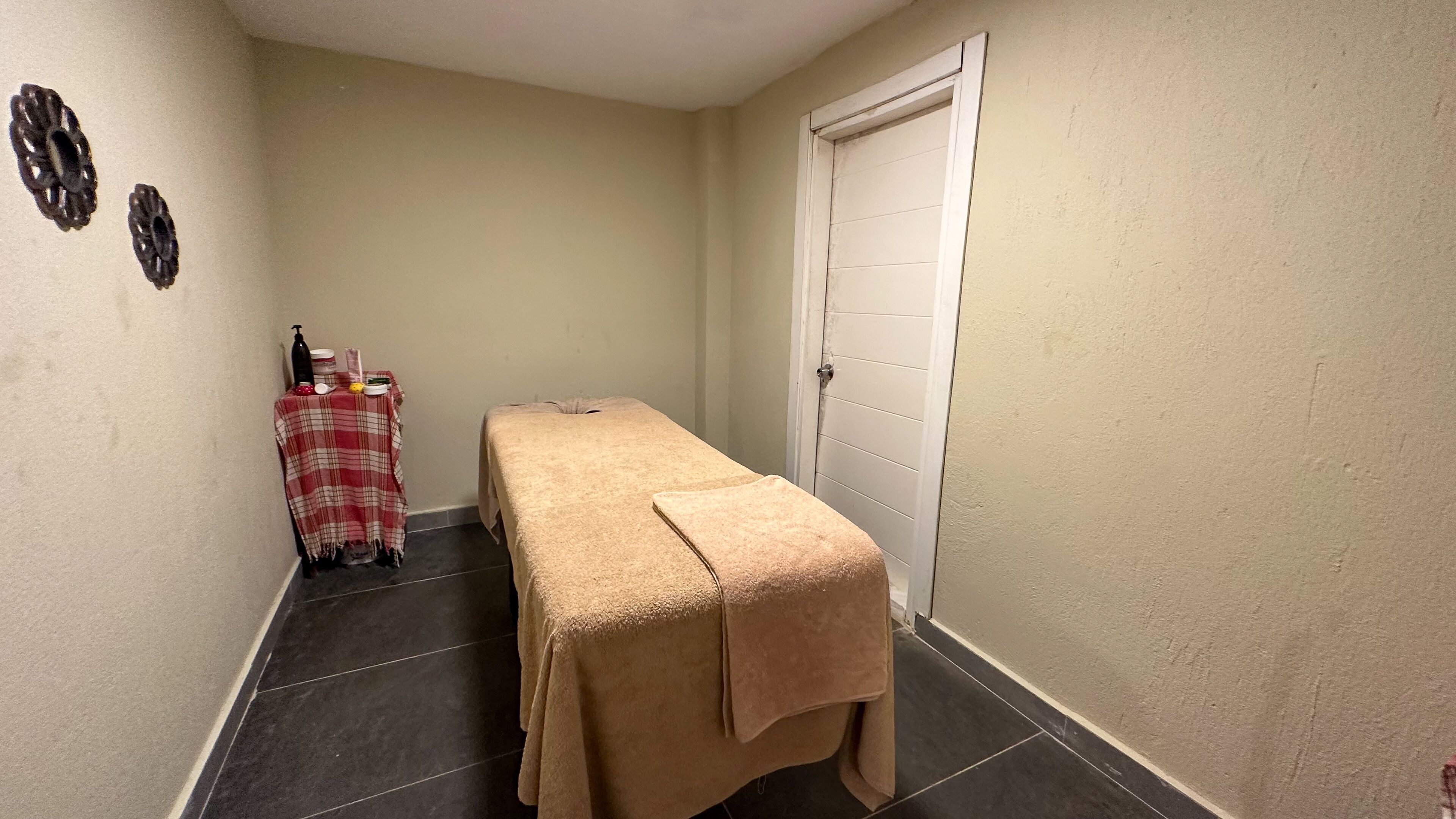 Um quarto pequeno e estreito com uma cama de solteiro coberta por um cobertor bege, uma mesa pequena com toalha xadrez vermelha e branca, e uma porta branca.