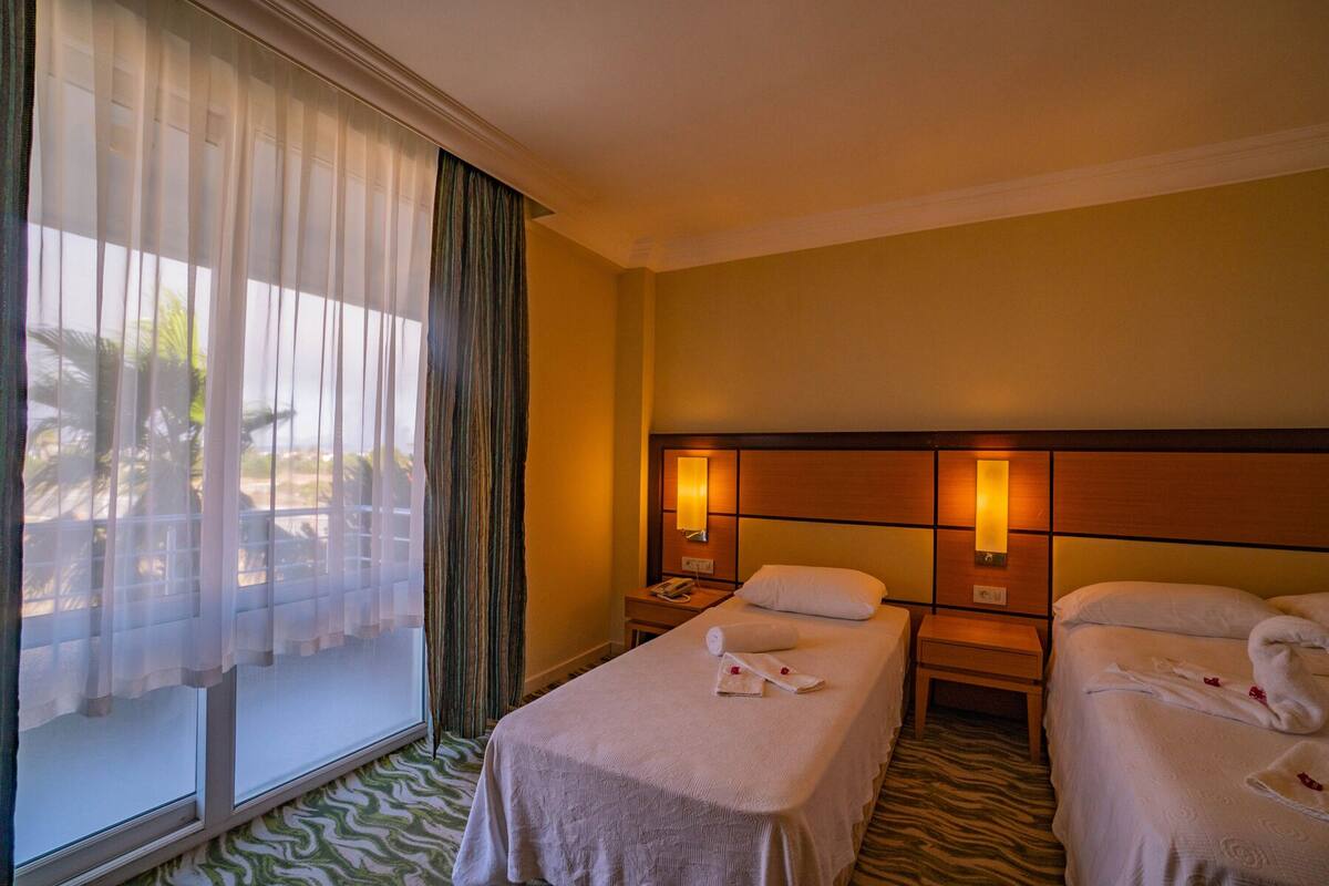 A hotel room with two beds, a large window with sheer curtains, and a view of palm trees.