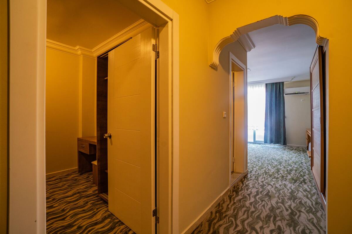 A hotel hallway with a patterned carpet, a closed door, and an arched doorway leading to a room with a window and curtains.