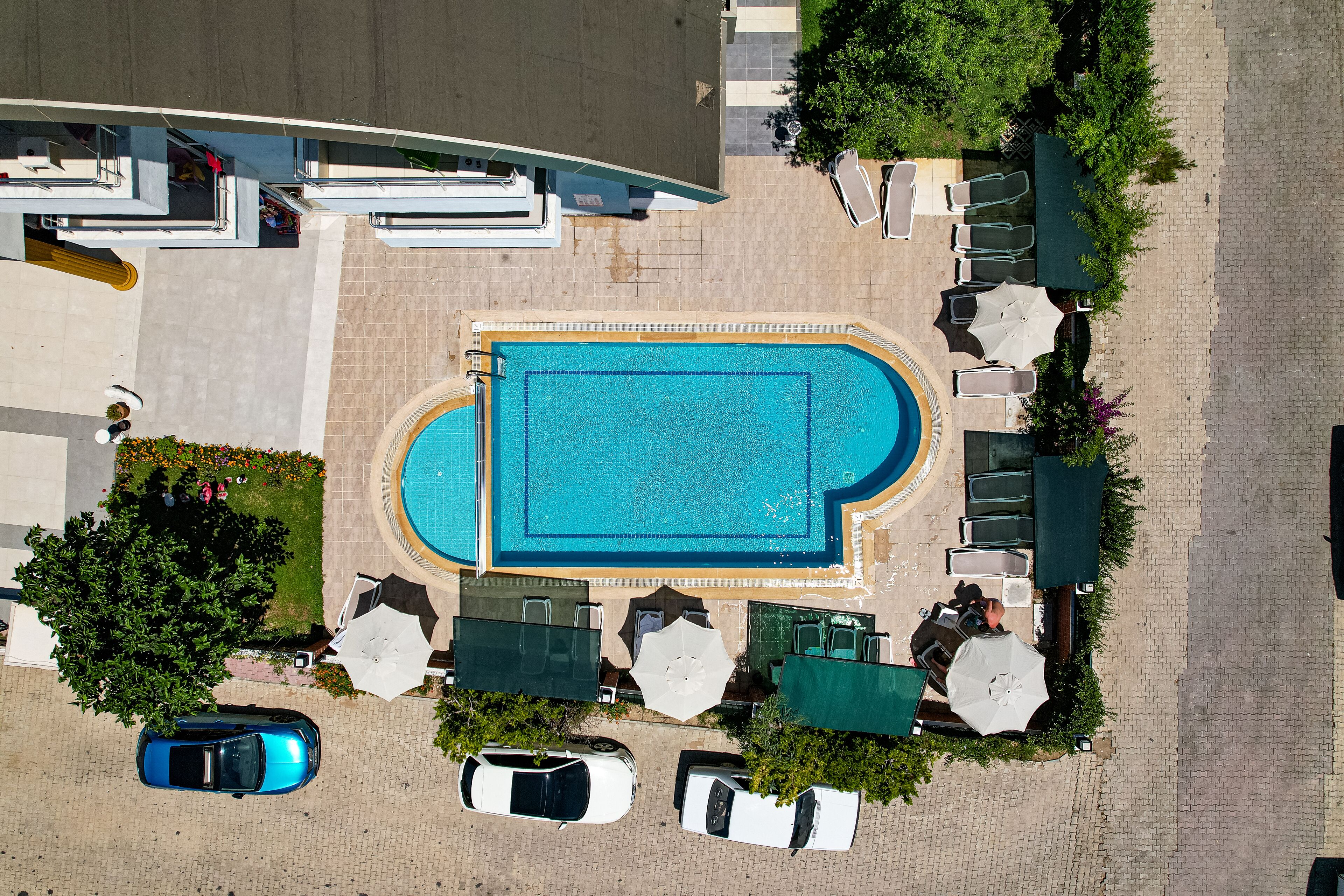An aerial view of a swimming pool area with lounge chairs, umbrellas, and a few cars parked nearby.