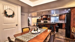 A spacious dining area with a wooden table, chairs, and a patterned runner. A kitchen with wooden cabinets and a refrigerator is visible in the background.