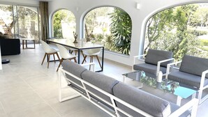Un salon moderne avec une table à manger en verre, des chaises blanches et des canapés gris. De grandes fenêtres offrent une vue sur la verdure et une structure en pierre à l’extérieur.