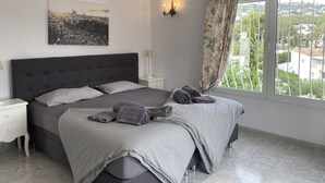 Une chambre à coucher avec un grand lit, une table de chevet, une lampe fixée au mur, une fenêtre avec des rideaux et un tableau représentant un paysage urbain.