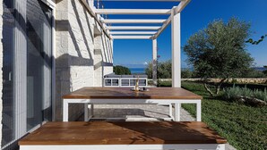 Un patio al aire libre con una mesa de madera y sillas blancas, con vistas a un jardín y un edificio de piedra frente al mar.