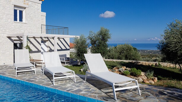 Zona junto a la piscina con sillones blancos, piscina de piedra y vistas al mar y al paisaje.
