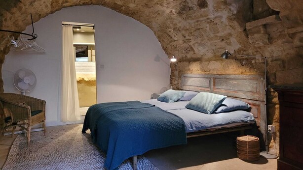 A bedroom with a stone wall, a bed with blue bedding, a wooden headboard, a fan, and a wicker chair. - Au Mûrier Blanc (Collias)