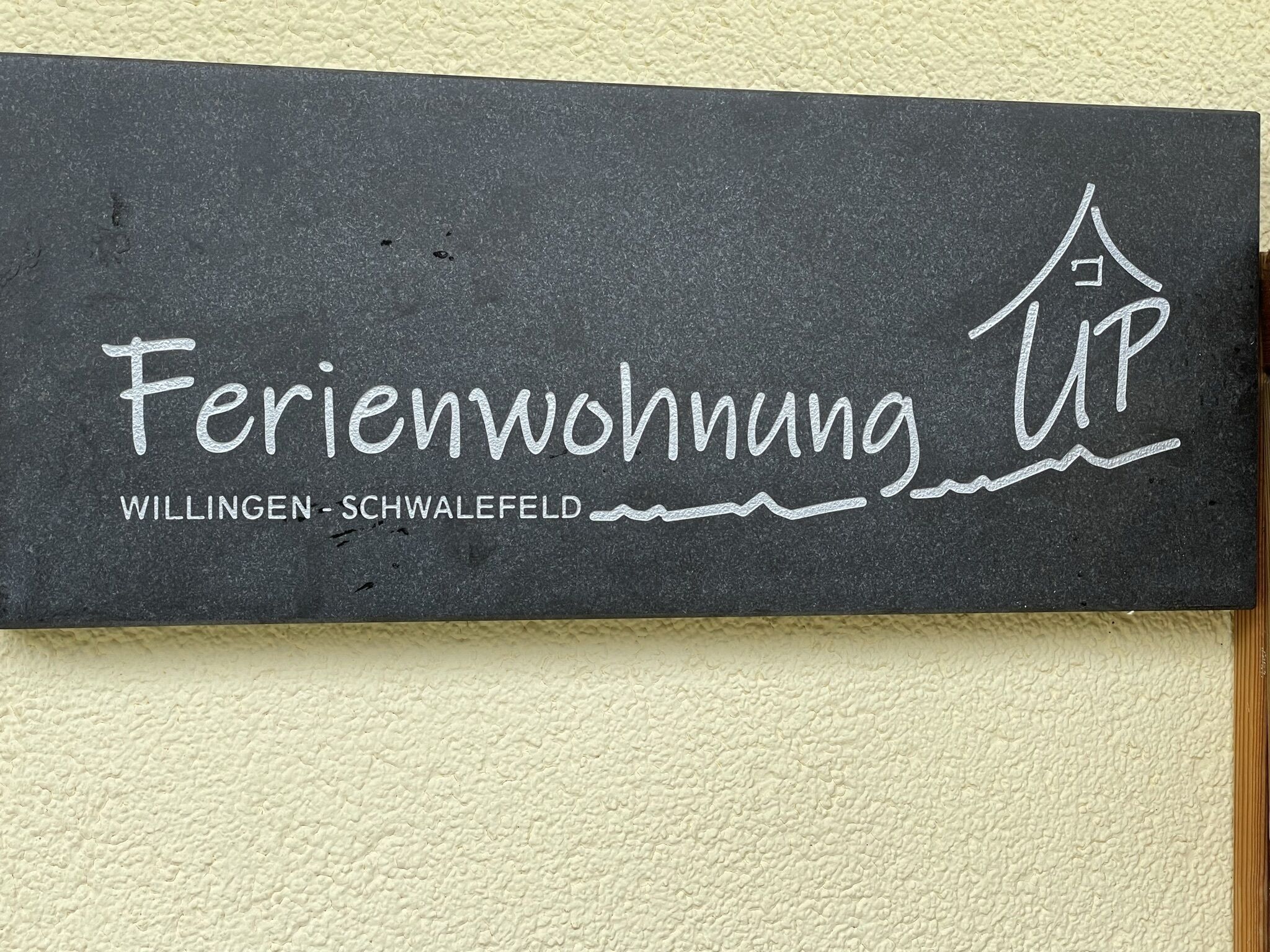 Un letrero con el texto 'Ferienwohnung UP WILLINGEN - SCHWALEFELD' y un sencillo icono de casa.